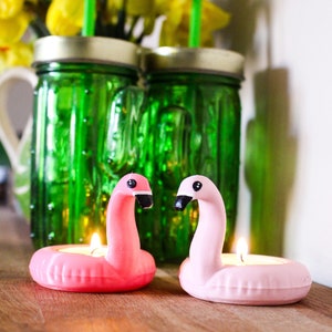 Può includere: Due portacandele a forma di fenicottero rosa con candele tealight accese. I portacandele sono su una superficie di legno davanti a due barattoli di vetro verde con cannucce.