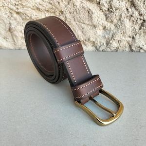 Peut inclure: Une ceinture en cuir marron enroulée avec une boucle en laiton. La ceinture présente des coutures blanches sur les bords et une boucle en cuir assortie. La boucle est de forme rectangulaire classique.