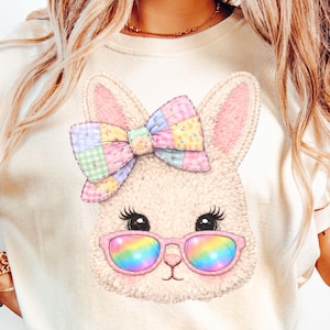 Puede incluir: Camiseta color crema con la cara de un conejito de dibujos animados con un lazo colorido y gafas de sol arcoíris. El conejito tiene orejas y pestañas rosas. La camiseta se combina con unos vaqueros azules.