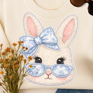 May include: Cream-colored sweatshirt featuring a bunny face with a blue floral bow and glasses. The bunny has pink inner ears and eyelashes. Sunglasses and a bouquet of dried flowers are also in the image. The sweatshirt is paired with blue jeans.