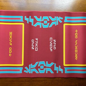 May include: A maroon rectangular mouse mat with yellow and teal detailing. The mat features teal and yellow stylized text and geometric designs. The text is in a unique font. The mat is on a wooden surface.