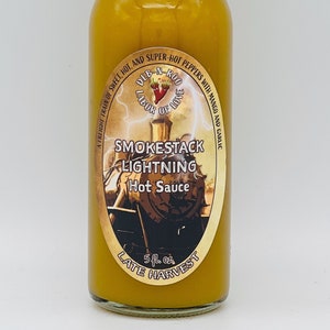 Puede incluir: Una botella de 5 fl oz de salsa picante Smokestack Lightning. La botella es amarilla y tiene una etiqueta con una ilustración vintage de una locomotora. La etiqueta también dice "Late Harvest".