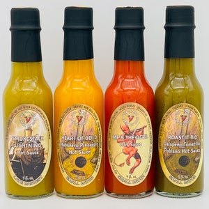 May include: Four small glass bottles of hot sauce with colorful labels. The labels feature illustrations and text, including the names "Smokestack Lightning", "Heart of Gold", "Me & The Devil", and "Roast It Big". Each bottle is 5 fl oz and labeled "Small Batch".