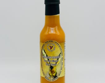 Op de afbeelding: Een doorzichtige glazen fles gevuld met oranje hete saus, afgesloten met een zwarte dop. Het etiket bevat een illustratie van een gele haas, de tekst "YELLOW RABBIT Carrot Ginger Hot Sauce", "DUB-N-KOO" en "5 FL OZ" in een cirkelvorm.