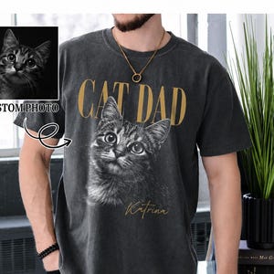 May include: A charcoal gray short-sleeve t-shirt featuring the gold text CAT DAD above a black and white portrait of a tabby cat. Below the cat, the name Katrina is printed in gold script. A small inset shows the original cat photo.