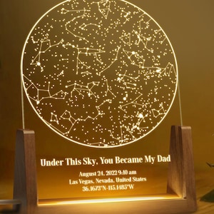 May include: A personalized night light featuring a circular design of a star map. The light is supported by a wooden base and displays the text "Under This Sky, You Became My Dad" along with a date, time, and location.