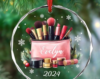 Op de afbeelding: Een heldere, ronde kerstbal met een rood lint. De ornament toont een roze make-uptas met de naam "Evelyn" en het jaar "2024". In de tas zitten make-up borstels en lippenstiften. Sneeuwvlokken zijn verspreid over het ontwerp.