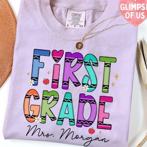 May include: A lilac Comfort Colors t-shirt with the words "FIRST GRADE" in colourful crayon-style letters. The shirt also features the name "Mrs. Morgan" in black script, along with small apple and flower graphics.