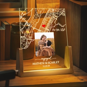 May include: A personalized illuminated map of New York, USA, with a red heart marking a location. The wooden base displays a photo of a couple and the text "engaged" with names and a date. The lamp is lit with a warm glow.