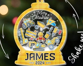 May include: A yellow and black Christmas ornament shaped like a snow globe, featuring construction vehicles and tools. The ornament has the name "JAMES" and the year "2024" printed on it, with the words "Merry Christmas" at the top.