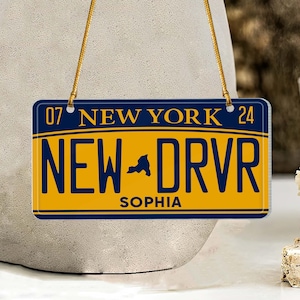 Puede incluir: Un adorno de placa de matr&iacute;cula de Nueva York amarillo y azul con el texto "NEW DRVR" y el nombre "SOPHIA" impreso en &eacute;l. La placa tiene los n&uacute;meros "07" y "24" a cada lado del texto "NEW YORK".