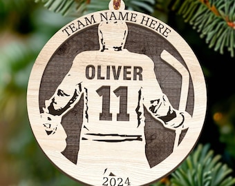 May include: A wooden hockey-themed Christmas ornament. The ornament features a hockey player silhouette with the name "OLIVER" and the number "11". The text "TEAM NAME HERE" and the year "2024" are also included.