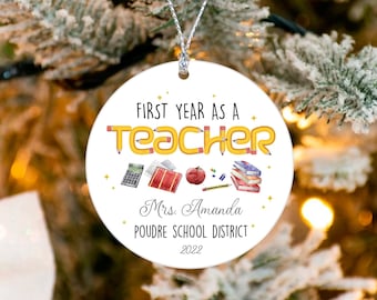 May include: A white, circular Christmas bauble with the text "FIRST YEAR AS A TEACHER" in black and yellow pencil-shaped letters. The bauble includes school-themed illustrations and the name "Mrs. Amanda" and "POUDRE SCHOOL DISTRICT 2022".