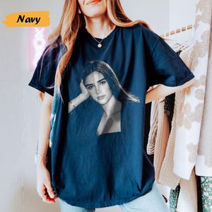 May include: Navy blue t-shirt featuring a black and white portrait of a woman. The word "Navy" is displayed in a yellow box in the upper left corner. The shirt is a solid color and has short sleeves.