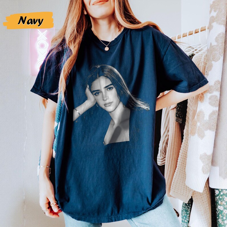 May include: Navy blue t-shirt featuring a black and white portrait of a woman. The word "Navy" is displayed in a yellow box in the upper left corner. The shirt is a solid color and has short sleeves.
