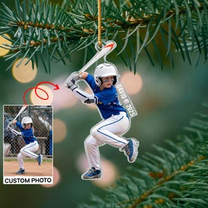 Puede incluir: Un adorno acrílico transparente que representa a un jugador de béisbol en posición de bateo, con uniforme azul y casco blanco. El adorno incluye el texto "DAVID 2025" y una imagen más pequeña con el texto "CUSTOM PHOTO".