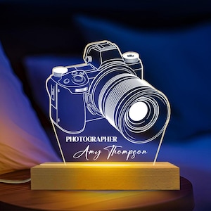 May include: A glowing acrylic camera night light with the word "PHOTOGRAPHER" and the name "Amy Thompson" etched on the base. The camera design is white and sits on a wooden base.