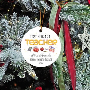 Puede incluir: Adorno navide&ntilde;o redondo y blanco con el texto "FIRST YEAR AS A TEACHER" en letras amarillas estilo l&aacute;piz. El adorno incluye ilustraciones escolares y el nombre "Mrs. Amanda" y "POUDRE SCHOOL DISTRICT 2022".