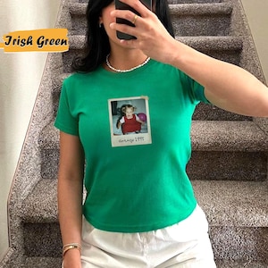Peut inclure: Un t-shirt vert avec une photo vintage d'une jeune fille tenant un ballon rose. La photo est dans un cadre blanc avec le texte "Vintage 1995" en dessous.