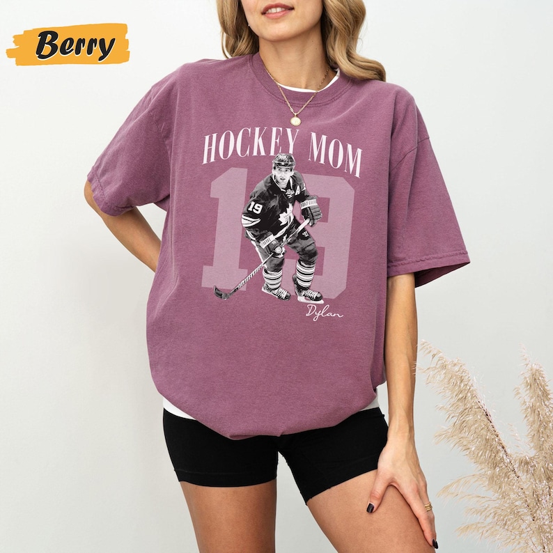Może przedstawiać: Koszulka w kolorze jagodowym z napisem "HOCKEY MOM" i grafiką hokeisty z numerem 19. Imię gracza, "Dylan", jest wydrukowane poniżej. Koszulka ma swobodny styl.