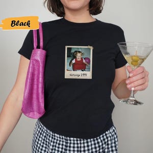 Peut inclure: T-shirt noir avec une photo vintage d'une jeune fille tenant un ballon rose. La photo est encadr&eacute;e d'une bordure blanche et porte le texte "vintage 1995" &eacute;crit en dessous.