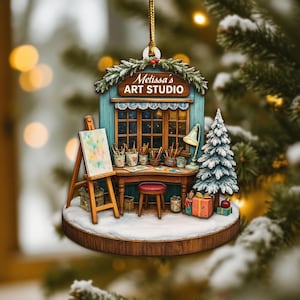 May include: A Christmas ornament featuring "Melissa's Art Studio." The ornament depicts a miniature art studio with a painting on an easel, a desk with art supplies, a small Christmas tree, and wrapped gifts. The studio is set on a snowy base.