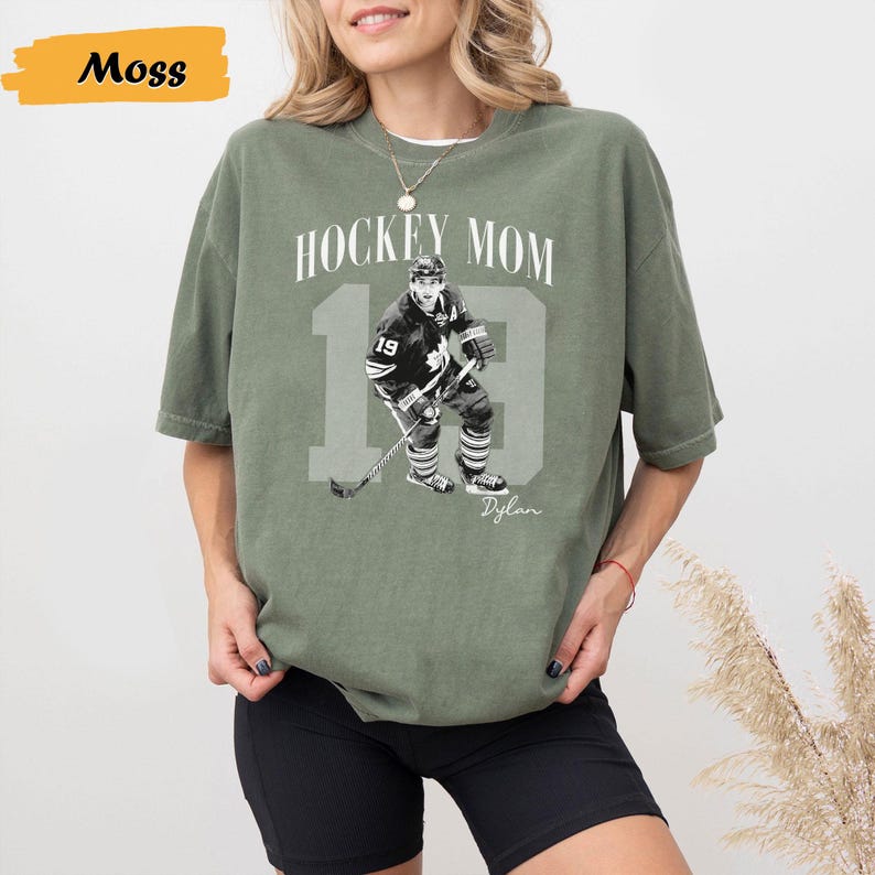 Może przedstawiać: Oliwkowa koszulka z napisem "HOCKEY MOM 19 Dylan" oraz czarno-białym zdjęciem hokeisty. Koszulka ma kr&oacute;tkie rękawy i okrągły dekolt.