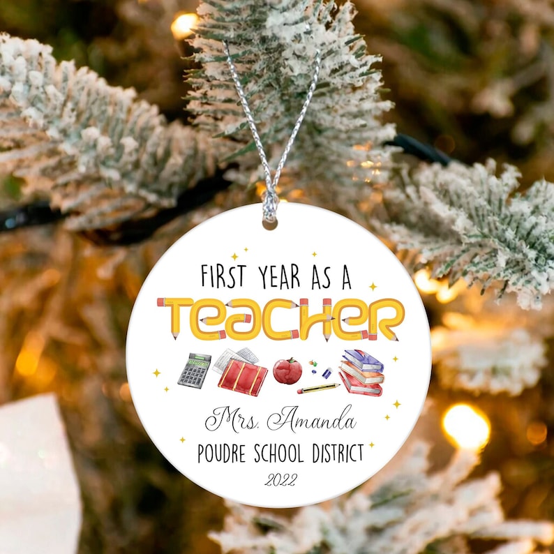 Puede incluir: Un adorno navide&ntilde;o redondo y blanco con el texto "FIRST YEAR AS A TEACHER" en letras negras y amarillas con forma de l&aacute;piz. El adorno incluye ilustraciones con tem&aacute;tica escolar y el nombre "Mrs. Amanda" y "POUDRE SCHOOL DISTRICT 2022".