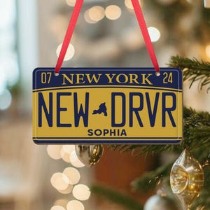 Puede incluir: Un adorno de placa de matr&iacute;cula de Nueva York amarillo y azul con el texto "NEW DRVR" y el nombre "SOPHIA" impreso en &eacute;l. El adorno est&aacute; colgado de una cinta roja.