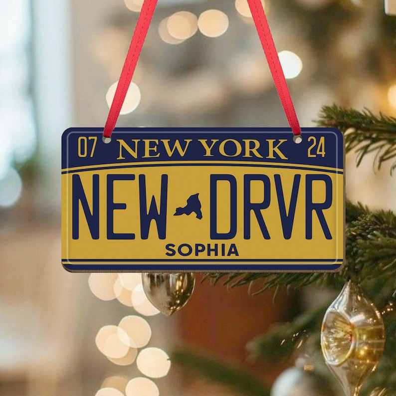Puede incluir: Un adorno de placa de matr&iacute;cula de Nueva York amarillo y azul con el texto "NEW DRVR" y el nombre "SOPHIA" impreso en &eacute;l. El adorno est&aacute; colgado de una cinta roja.