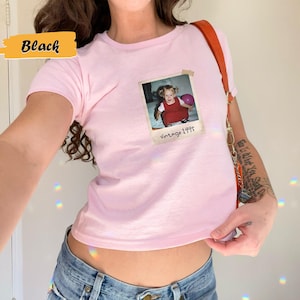Peut inclure: Un t-shirt rose avec une photo vintage d'une jeune fille tenant un ballon. La photo est encadr&eacute;e d'une bordure blanche et porte le texte "Vintage 1995" en dessous.