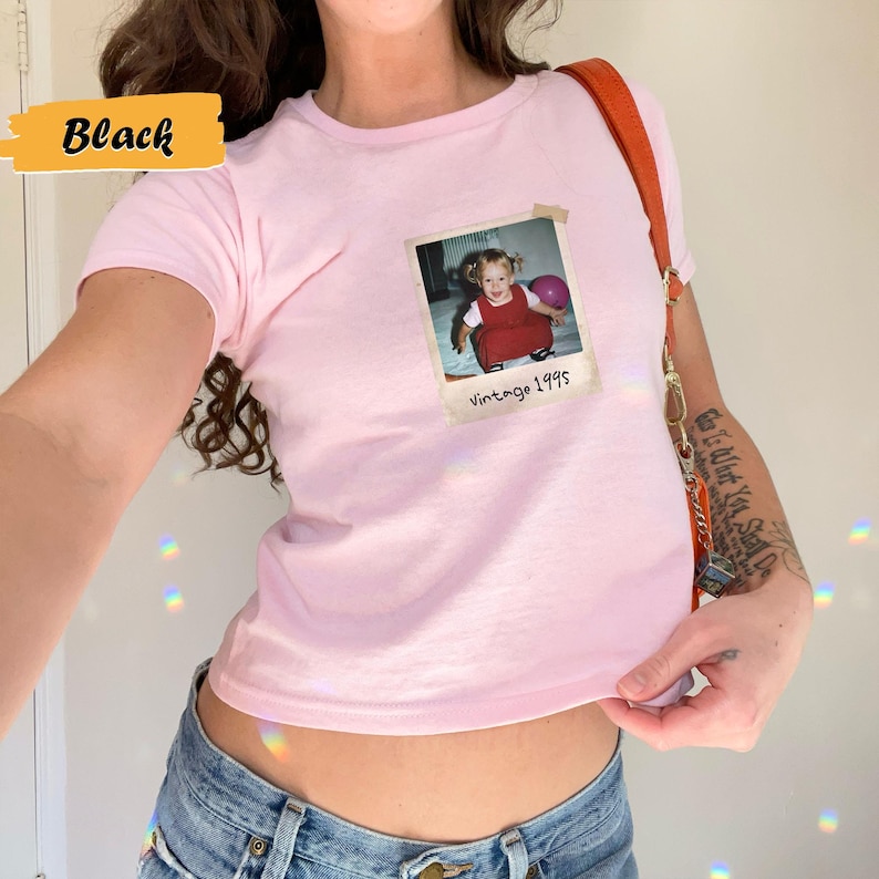 Peut inclure: Un t-shirt rose avec une photo vintage d'une jeune fille tenant un ballon. La photo est encadr&eacute;e d'une bordure blanche et porte le texte "Vintage 1995" en dessous.