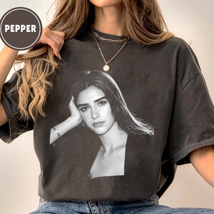 May include: Dark gray t-shirt featuring a black and white portrait of a woman. The shirt has a crew neck and short sleeves. The word "PEPPER" is printed on a circular label in the upper left corner.