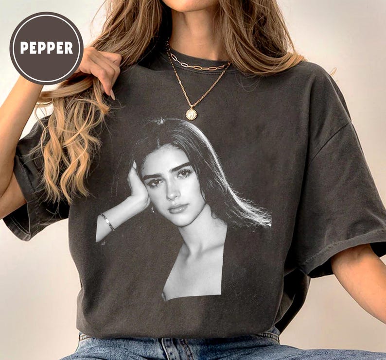 May include: Dark gray t-shirt featuring a black and white portrait of a woman. The shirt has a crew neck and short sleeves. The word "PEPPER" is printed on a circular label in the upper left corner.