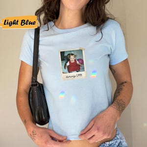 Peut inclure: T-shirt bleu clair avec une photo vintage d'une jeune fille tenant un ballon rose. La photo est dans un cadre blanc avec le texte "Vintage 1995".