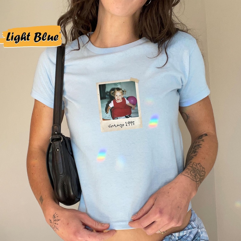 Peut inclure: T-shirt bleu clair avec une photo vintage d'une jeune fille tenant un ballon rose. La photo est dans un cadre blanc avec le texte "Vintage 1995".