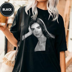 May include: Black oversized t-shirt featuring a black and white portrait of a woman. The shirt has a crew neck and short sleeves. The word "BLACK" is printed on a black circle on the upper left side of the image.