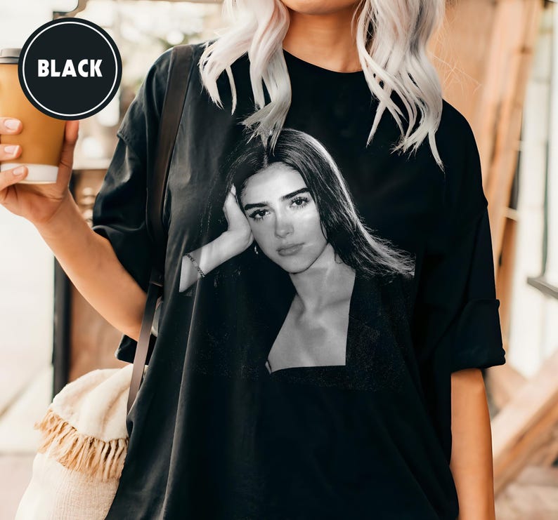 May include: Black oversized t-shirt featuring a black and white portrait of a woman. The shirt has a crew neck and short sleeves. The word "BLACK" is printed on a black circle on the upper left side of the image.