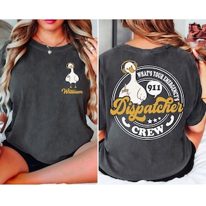 May include: A dark gray t-shirt with a white cartoon goose wearing headphones and holding a cup of coffee. The back of the shirt has a white circle with the text "What's Your Emergency's 911 Dispatcher Crew" in a yellow and white font.