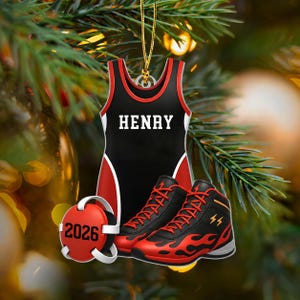 May include: A hanging ornament shaped like a black and red wrestling singlet with the name HENRY printed in white, paired with a pair of black and red wrestling shoes featuring flame patterns and a red circular headgear piece marked with 2026.