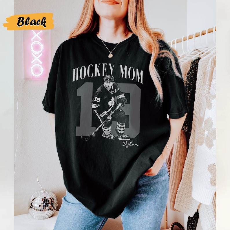 Może przedstawiać: Czarna koszulka z napisem "HOCKEY MOM" nad dużą liczbą 13 i wizerunkiem hokeisty. Zawodnik ma na sobie str&oacute;j hokejowy z numerem 19. Pod obrazkiem widnieje imię "Dylan".