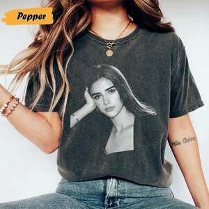 May include: Dark grey t-shirt featuring a black and white portrait of a woman. The shirt has a crew neck and short sleeves. The word "Pepper" is in a yellow rectangle in the upper left corner.