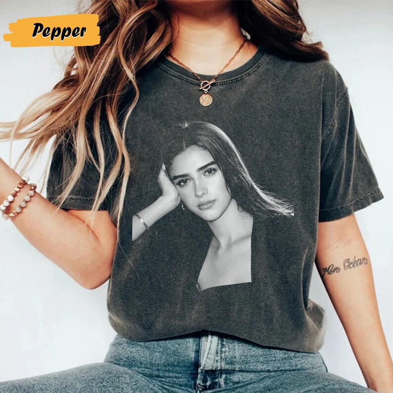 May include: Dark grey t-shirt featuring a black and white portrait of a woman. The shirt has a crew neck and short sleeves. The word "Pepper" is in a yellow rectangle in the upper left corner.