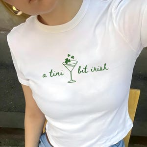 May include: White t-shirt with a green graphic of a martini glass and shamrocks. The text "a tini bit irish" is printed in green. The shirt has a crew neck and short sleeves.