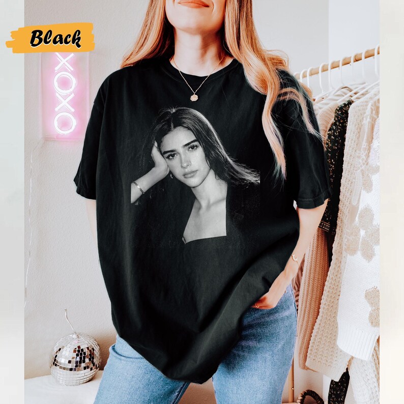 May include: Black t-shirt featuring a black and white portrait of a person. The t-shirt has a square neckline and short sleeves. The word "Black" is in a yellow box in the upper left corner. The word "OXOX" is in a pink neon sign.