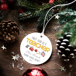Puede incluir: Un adorno navide&ntilde;o redondo blanco con el texto "First Year as a Teacher" en amarillo y naranja. El adorno incluye ilustraciones escolares y el nombre "Mrs. Amanda" y "Poudre School District 2022".