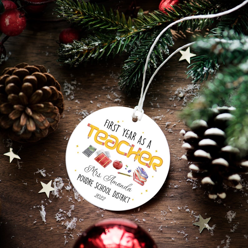 Puede incluir: Un adorno navide&ntilde;o redondo blanco con el texto "First Year as a Teacher" en amarillo y naranja. El adorno incluye ilustraciones escolares y el nombre "Mrs. Amanda" y "Poudre School District 2022".