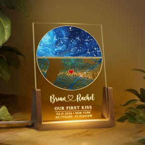 May include: A personalised night light with a celestial map and a city map, featuring the names "Brian & Rachel" and the text "OUR FIRST KISS". The date and coordinates are also displayed. The light sits on a wooden base.
