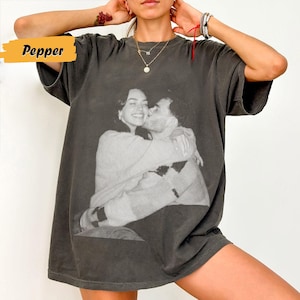 May include: Dark gray t-shirt featuring a black and white image of a couple embracing and kissing. The word "Pepper" is in a yellow box in the upper left corner. The shirt has short sleeves and a relaxed fit.