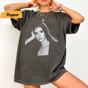 May include: Dark grey t-shirt featuring a black and white portrait of a woman. The word "Pepper" is in a yellow box on the upper left. The shirt has short sleeves and a relaxed fit, suitable for casual wear.